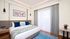 A well-appointed hotel bedroom features a large bed with blue and patterned cushions and a decorative throw, a blue rug on the wooden floor, and a small round table. - Iris Sarovar Portico, Sector 85, Gurugram