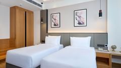 A comfortable hotel room is furnished with two single beds with white linens, a wooden wardrobe, a bedside table, and framed artwork on the wall above the headboards. - Iris Sarovar Portico, Sector 85, Gurugram