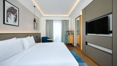 A bright hotel room is furnished with a large bed with white linens, a blue rug, a wall-mounted TV, a mini-fridge unit, and a chair by the window with grey curtains. - Iris Sarovar Portico, Sector 85, Gurugram