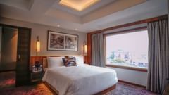  Room at Ishaan Sarovar Portico, Deoghar featuring a bed, a bedside table with lamps on either side, and a large window with curtains.