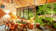 An image showing wooden chairs and table placed aesthetically inside a room with bamboo roofing, where the garden view is at an enjoyable distance