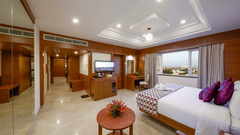 A well-lit suite at JP Hotel Chennai featuring a plush bed next to a centre table facing the TV  and a large window on its side