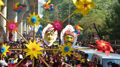 A procession on a road with DIY fans in the foreground and people carrying busts on a pole next to parked cars during Poila Boishakh.