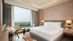 A bedroom at KPG Sarovar Portico, Raj Nagar Extension, Ghaziabad featuring a king size bed, a desk with chair, a window with outdoor views, and bedside tables with lamps.