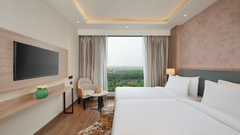 A bedroom at KPG Sarovar Portico, Raj Nagar Extension, Ghaziabad featuring twin beds, a wall mounted television, an armchair, and a window overlooking green fields.
