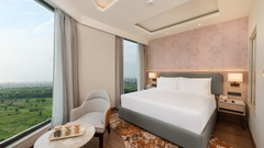 A bedroom interior at KPG Sarovar Portico, Raj Nagar Extension, Ghaziabad featuring a king size bed, bedside tables with lamps, an armchair, and floor-to-ceiling windows.