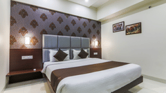A well-lit hotel room featuring a large neatly made double bed with brown accents, padded headboard, patterned wallpaper, bedside lamps, and framed artwork on a light-coloured wall - Magnus Square, Viman Nagar.