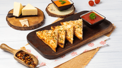 A wooden board holds triangular pastries next to cheese wedges, bowls of sauce, and a scoop of chilli flakes on a wooden surface.