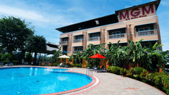 Facade of MGM Vailankanni Residency featuring a large swimming pool on a sunny day surrounded by lawns and bushes