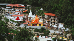 A scenic view of the colourful Kainchi Dham with red and white domes nestled in a lush green valley by a river.
