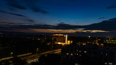 An aerial view of a city at dusk, showing street lights and buildings, with a prominent multi-storey building in the middle distance against a dark blue and orange sky