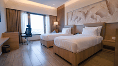 A bright hotel room featuring twin beds, a wall-mounted piece of art above the beds, and a large window offering outdoor views | Pride Elite Chhatrapati Sambhajinagar