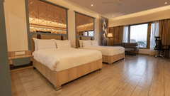 A well-lit hotel room featuring twin beds, a sofa near a window, wooden flooring, and a patterned headboard or wall panel | Pride Elite Chhatrapati Sambhajinagar