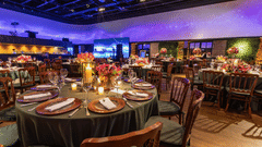 A wide shot of a large indoor venue set up for a formal event or gala dinner, with numerous round tables, chairs, and ambient lighting.