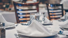 A close-up of a neatly set dining table with white tablecloth, a folded white napkin, a wine glass, and cutlery.