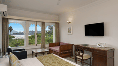 A guest room at Ram Pratap Palace with a bed, pillows, two chairs, a coffee table, a desk, a television, a lamp, and a window with arched frames overlooking the lake.