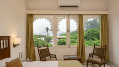 A guest room at Ram Pratap Palace in Udaipur, with a bed, two chairs, a side table, and a window with arched frames overlooking the outdoors.