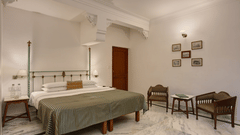 A  bedroom at Ram Pratap Palace, Udaipur, featuring a well made four-poster bed, two wooden chairs, a small table, and framed art on the wall.