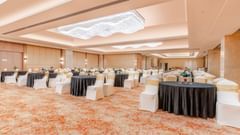 A ballroom at Sarovar Premiere Alwar with round tables and chairs arranged on a patterned carpet floor.