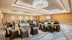 A conference room at Sarovar Premiere Alwar with round tables and chairs with gold sashes and a projector screen.