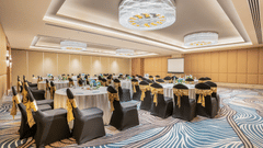 A conference room at Sarovar Premiere Alwar with round tables and chairs with gold sashes and a projector screen.