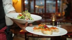 A server holds two white plates, one with a salad and the other with bruschetta, in a restaurant setting.