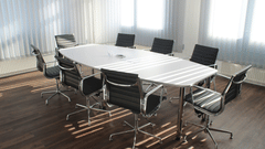 A bright, empty modern boardroom with a large white oval table and black rolling office chairs.