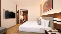 Skywings Sarovar Portico Mathura deluxe bed room with wall-mounted TV, wooden wardrobe, gray sofa, red framed wall art, and herringbone wood flooring.