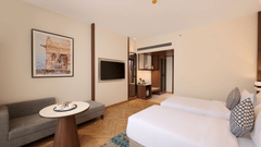 Skywings Sarovar Portico Mathura twin bed room with sofa seating, round coffee table, wall art, TV unit, and warm wood flooring.