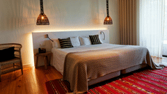 A modern guest room with a bed dressed in a striped grey throw and cushions, woven pendant lights over a backlit headboard, and a rattan armchair.