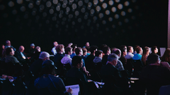 Audience seated in dimly lit room, listening attentively. A backdrop of circular lights adds ambiance. The mood is focused and engaged.