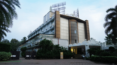 Facade of Sarovar Portico, Bhubaneswar, seen under a bright and partially cloudy sky, with a well-lit driveway, a manicured garden, and a board with the name of the hotel on it.