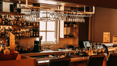 A commercial bar area with many bottles on shelves, stemware hanging above the counter, and seating stools below.