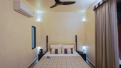 A well-lit photograph showing the elegant simplicity of the Sierra Jacuzzi Suite's bedroom. The comfortable four-poster bed, dressed in crisp white and red bedding, is centered between two bedside tables under a high ceiling with a ceiling fan.