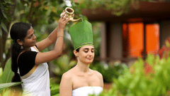 A woman in traditional attire pours herbal oil onto a relaxed guest’s head, covered with a green leaf wrap, in a serene setting at Uday Backwater Resort