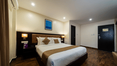 A clean, modern bedroom at VITS Select, Bengaluru, featuring a large bed, shining wooden flooring, a prominent headboard, and a closed door leading into the room.