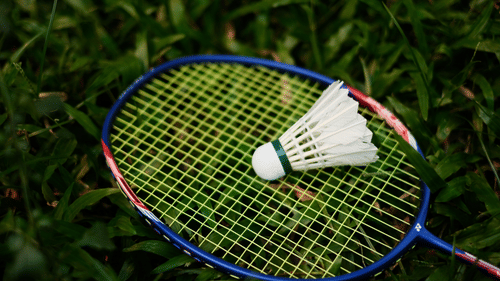 A single white shuttlecock resting on the yellow strings of a badminton racket lying on green grass.