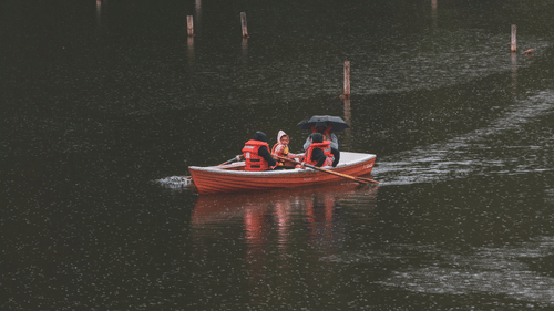 A small orange rowing boat carrying 3 people gliding across the calm surface of a dark lake.