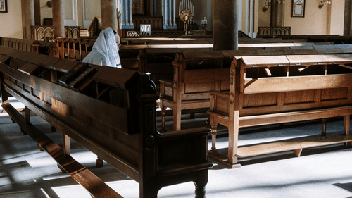 A person in prayer sits alone on a wooden pew inside a quiet, sunlit church with tall columns and an altar.