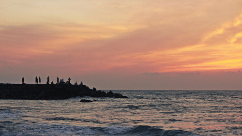 A vibrant orange and pink sunset over the ocean, with a silhouetted breakwater and figures in the distance.