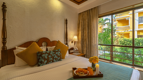 An image of a room showcasing a beautifully decorated bed adorned with a tray of candles, flowers, and fruits, complemented by a large window.