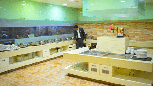 A staff member stands near a long buffet counter area at Ambar Sarovar Portico, Gandhidham equipped with metal food warmers and clean plates.