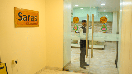 An entrance to Saras restaurant at Ambar Sarovar Portico, Gandhidham features a glass door, a staff member, and a wooden signboard on the wall.