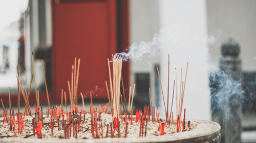 A shallow-focus shot of an incense bowl with many burning sticks and wisps of smoke rising into the air.