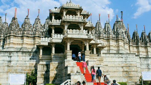 A beautiful white marble Jain temple with many smaller spires. A red carpet leads up the steps to the main entrance.