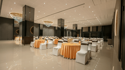 A spacious event hall with numerous round tables and chairs, set for a gathering, under soft lighting - Biznotel By Pride Surat