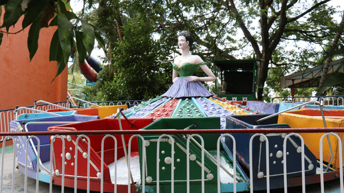 A colourful, vintage carousel ride with a mannequin in a green and purple dress at the centre, surrounded by trees and a fence at Black Thunder, Coimbatore.