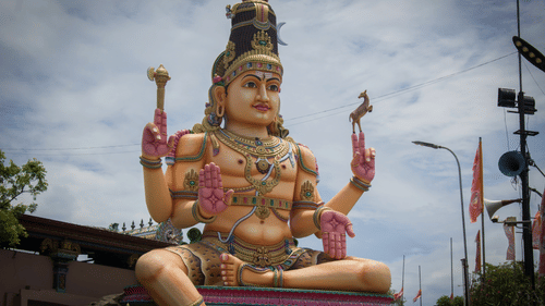A towering statue of an idol in a seated pose, adorned with traditional ornaments and holding symbolic objects, set against a cloudy sky.