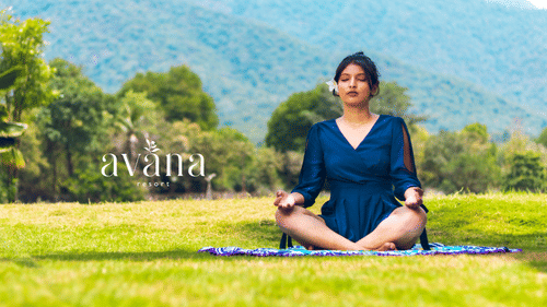 Woman practicing meditation on a yoga mat in a lush green field with scenic mountain views.