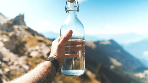 A person holding a reusable glass water bottle amidst mountains.